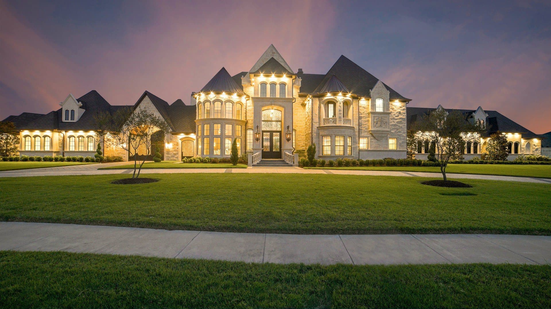 A stone estate home glowing at dusk across a manicured lawn